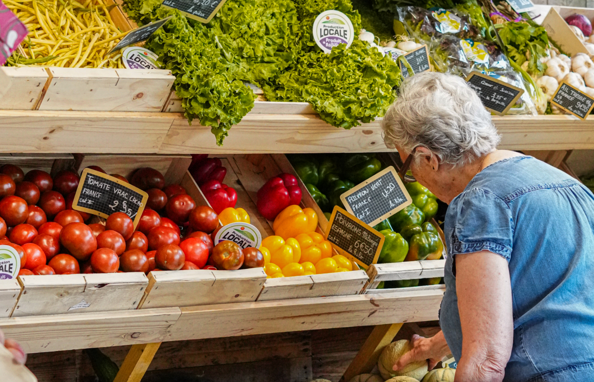 Une vieille dame de dos se penche devant un étalage de fruits et légumes frais colorés